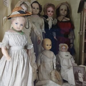 Collection of antique porcelain dolls in Victorian dresses displayed in a cabinet, including a large girl doll in a floral dress with a straw hat.