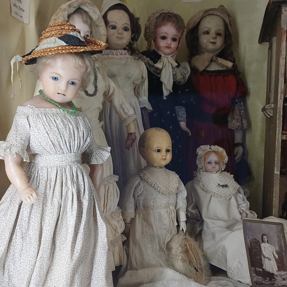 Collection of antique porcelain dolls in Victorian dresses displayed in a cabinet, including a large girl doll in a floral dress with a straw hat.