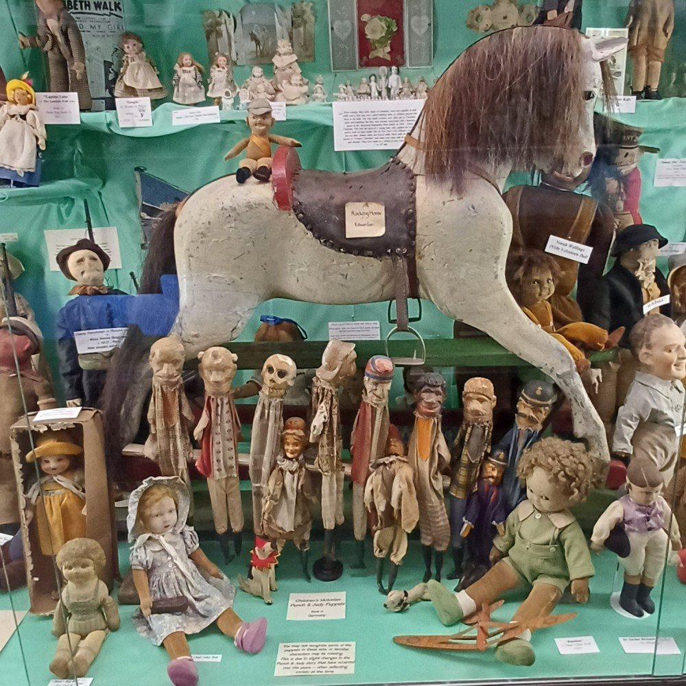 Antique toy display in a glass case, centered on a white rocking horse with a brown saddle, surrounded by dolls and puppets.